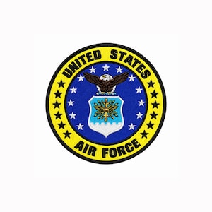 Puede incluir: Un parche bordado circular con el emblema de la Fuerza Aérea de los Estados Unidos. El diseño incluye un escudo central con un elemento heráldico dorado sobre un fondo azul claro, coronado por un águila marrón, rodeado de estrellas blancas.