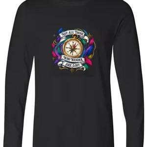 May include: A black long-sleeve shirt featuring a central graphic of a gold compass surrounded by colorful feathers and a white banner with the text "NOT ALL THOSE WHO WANDER ARE LOST" printed in black capital letters across the front chest area.