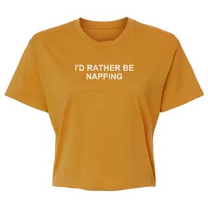 May include: A mustard yellow short-sleeved t-shirt with a crew neckline. The center of the chest features the white text "I'D RATHER BE NAPPING" printed in a clean, sans-serif font. The garment has a straight hemline and a simple, casual design.