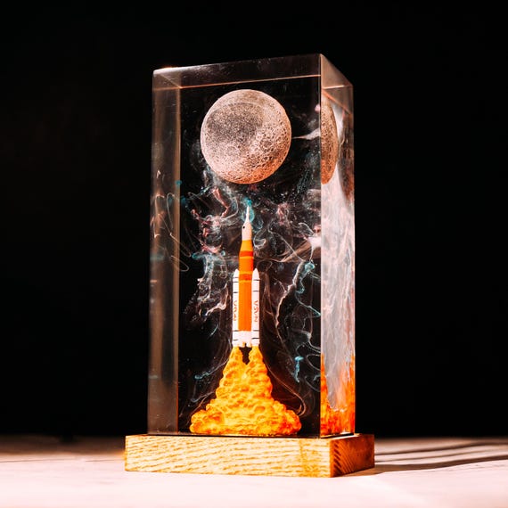 A handmade resin diorama of the Artemis II rocket launch — real mission, real craft, glowing LED galaxy trapped in resin that doubles as a night light and a nerdy heirloom.