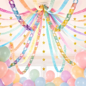 Puede incluir: Una decoración de techo con serpentinas de papel pastel y cadenas de papel entrelazadas en tonos rosa, amarillo, azul y morado. Guirnaldas de estrellas doradas cuelgan verticalmente entre las serpentinas, junto a globos pastel.