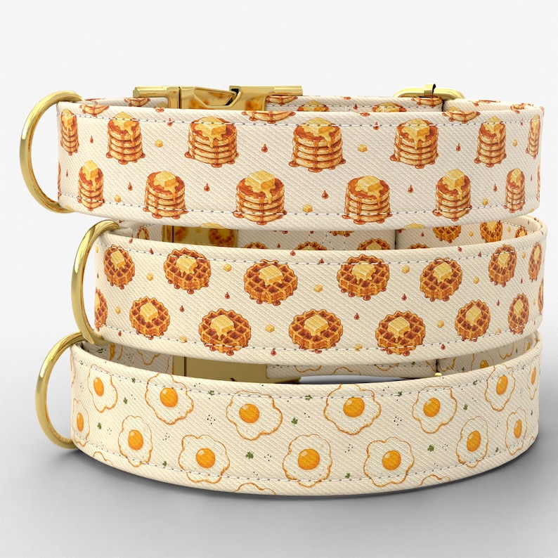 Puede incluir: Tres collares para mascotas apilados verticalmente, cada uno con una correa de tela color crema con un estampado de comida repetitivo. El collar superior muestra pilas de panqueques, el medio gofres y el inferior huevos fritos, con herrajes dorados.