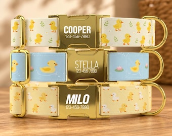 May include: Three pet collars with gold-tone metal buckles and D-rings. The top collar is cream with yellow ducks, the middle is light blue with ducks and lily pads, and the bottom is yellow with white and yellow ducks. Each buckle displays a name and phone number.