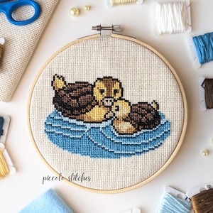 May include: A cross-stitch embroidery hoop with a design of two brown and yellow ducks swimming in blue water. The hoop is surrounded by various colors of embroidery floss.