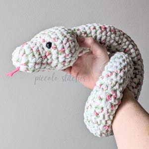 May include: A plush, crocheted snake in white with pink and green accents. The snake has a black eye and a pink tongue. The text "piccolo stitches" is visible on the image.