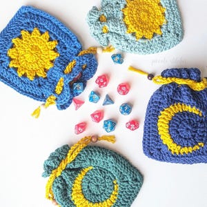 May include: Crocheted dice bags in blue, teal, and yellow, featuring sun and moon designs. The bags are square-shaped with drawstring closures and wooden beads. Scattered around are various colorful polyhedral dice.