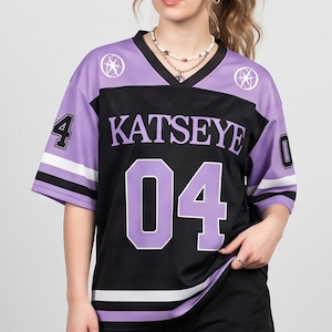 Puede incluir: Una camiseta de manga corta con un panel central negro que presenta el texto KATSEYE y el número 04 en una fuente morada grande. Las mangas y la parte superior del pecho son de color morado claro con rayas blancas y negras. Dos logotipos circulares de estrellas están en los hombros.