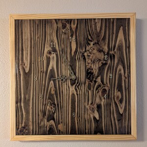 Puede incluir: Un reloj de pared cuadrado con una esfera de madera oscura y quemada con nudos y texturas naturales visibles. El reloj tiene un marco de madera clara y manecillas de metal plateado. Los números del 1 al 12 aparecen en una fuente plateada.