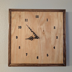 Puede incluir: Un reloj de pared cuadrado con una esfera de madera clara de grano natural enmarcada por un borde de madera más oscura. El reloj tiene números arábigos negros del 1 al 12 y manecillas de metal negro ornamentadas en el centro.