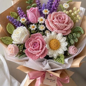 May include: A crochet flower bouquet featuring pink and white roses, daisies, and purple lavender sprigs with green leaves. The arrangement is wrapped in brown paper and white tissue, tied with a pink ribbon and a tag reading Handmade with love.
