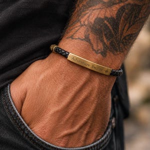 Op de afbeelding: Een zwarte gevlochten leren armband met een rechthoekige, messingkleurige metalen plaat. De plaat is gegraveerd met de tekst "Adventure Awaits" en een klein kompasroossymbool. De armband heeft een metalen sluiting en een getextureerd koordontwerp.