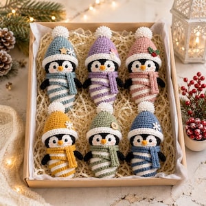 Könnte beinhalten: Ein Set aus sechs gehäkelten Pinguin-Ornamenten in einer Pappschachtel. Jeder Pinguin trägt eine bunte Strickmütze mit weißem Bommel und einen passenden Schal. Die Pinguine haben schwarz-weiße Körper mit orangefarbenen Schnäbeln und kleinen Augen.