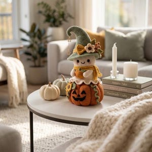 May include: A crochet doll featuring a pale face and a green pointed hat adorned with small flowers and a dangling star charm. The doll wears a yellow scarf and sits atop a large orange jack-o'-lantern with a carved face and green accents.
