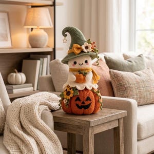 May include: A crocheted ghost figurine wearing a green pointed hat with a small star charm, a yellow scarf, and holding a tiny orange pumpkin. The ghost sits atop a larger orange crocheted pumpkin decorated with sunflowers and green vines.