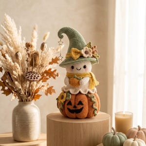 May include: A crocheted ghost figure wearing a sage green pointed hat with a yellow bow and flower, a yellow scarf, and holding a small orange pumpkin. The ghost sits atop a larger orange jack-o'-lantern with a carved face and green vines.