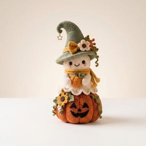 May include: A crocheted ghost figure wearing a sage green pointed hat with a small star charm, a yellow scarf, and holding a tiny orange pumpkin. The ghost sits atop a larger orange crocheted pumpkin with a black jack-o'-lantern face and sunflowers.