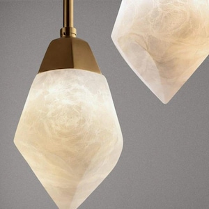 Puede incluir: Dos lámparas colgantes con pantallas geométricas en forma de diamante hechas de un material blanco translúcido con aspecto marmolado. Cada pantalla está unida a una tapa metálica dorada y un cable fino, emitiendo una luz suave y difusa.