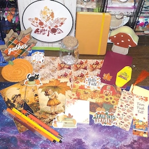 May include: A collection of autumn-themed stationery and decor including a yellow notebook, a glass jar, a mushroom-shaped block, a crochet pumpkin, stickers with text like Hello Fall and Fall Into Reading, bookmarks, and cards with vintage illustrations.