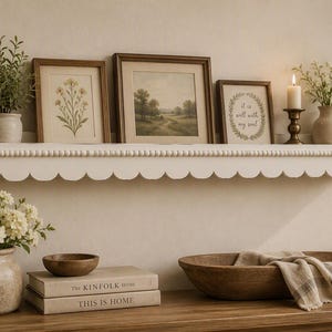 May include: A white wooden wall shelf with a scalloped edge and beaded trim detail. The shelf holds two framed botanical prints, a framed landscape painting, a framed quote reading "it is well with my soul," a lit candle, and a small potted plant.