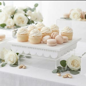 May include: A white scalloped cake stand made of wood or composite material, elevated on a clear acrylic base. The stand holds several vanilla cupcakes with white frosting and light beige macarons, surrounded by white roses and eucalyptus leaves.