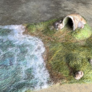 May include: A needle-felted landscape mat featuring a textured green grassy field with brown earth patches, a small arched cave structure, and a blue and white water section representing a shoreline with white foam waves.