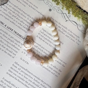 May include: A beaded bracelet featuring a seashell charm, alternating pink and white beads, and gold-colored spacers. The bracelet rests on a page of a book with text about spells and relationships.