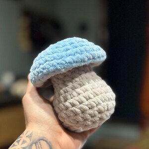 May include: A crocheted mushroom toy, featuring a light blue cap and a beige stem. This soft, textured mushroom is a whimsical decorative item, suitable for nurseries or as a unique gift.