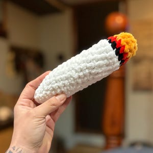 May include: A handmade, plush crochet item shaped like a lit cigarette. The body is white, textured yarn, while the tip features orange, yellow, red, and black yarn, mimicking a burning end. The item is held in a hand.