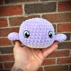 May include: A handmade, lavender crocheted whale with small flippers and large, cartoon-style eyes. The whale has a white belly and is held in a person's hand. The background is a brick wall.