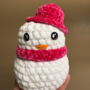 May include: A handmade snowman plushie with a white body, a bright pink hat, and a matching scarf. The snowman has black eyes and an orange carrot nose. The plushie is made of soft, textured yarn.