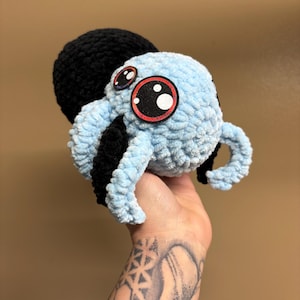 May include: A handmade, light blue crocheted octopus plush toy with large, cartoonish eyes outlined in red and black. The octopus has a black head and tentacles. The toy is soft and fluffy, and appears to be held in a hand.