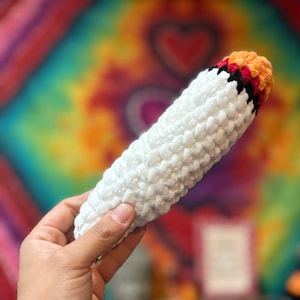 May include: A plush, cylindrical crochet toy featuring a white body, a black band, and a textured orange tip. The item is crafted from soft, chunky yarn with a visible stitch pattern throughout, creating a tactile, rounded, and elongated shape.