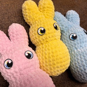 May include: Three plush bunny toys in pastel colors: pink, yellow, and blue. Each bunny has large, cartoonish eyes with black pupils and white and yellow accents. The bunnies are made of a soft, textured material.