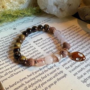 May include: A bracelet with a variety of natural stone beads in brown, gold, and pink hues. The bracelet, which includes a charm, is presented on a book with printed text.