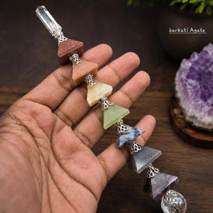 Puede incluir: Un adorno colgante vertical con una punta de cuarzo transparente en la parte superior, seguida de una serie de siete piedras triangulares en rojo, naranja, amarillo, verde, azul, gris y morado, terminando con una esfera transparente. Tapas de filigrana plateada conectan cada pieza.