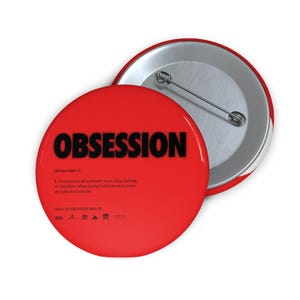Könnte beinhalten: Ein runder roter Ansteckbutton mit dem Wort OBSESSION in fetter schwarzer Schrift, gefolgt von einer wörterbuchartigen Definition, dem Text ONLY IN THEATERS MAY 13 und mehreren kleinen Logos am unteren Rand. Die Rückseite zeigt eine Metallnadel.
