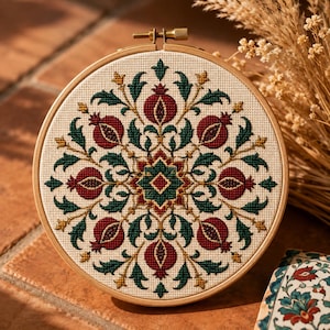 May include: A circular wooden embroidery hoop containing a cross-stitch pattern on cream-colored fabric. The design features a symmetrical arrangement of deep red pomegranates, dark green leaves, and gold accents radiating from a central star.