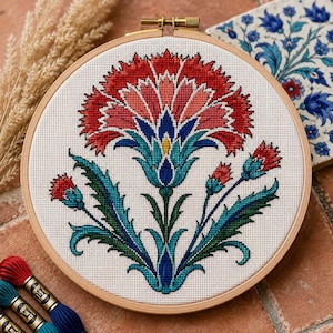May include: A circular wooden embroidery hoop containing a cross-stitch pattern of a large, stylized carnation flower. The design features vibrant red, pink, teal, and dark blue threads on a white fabric background, showcasing intricate floral motifs.