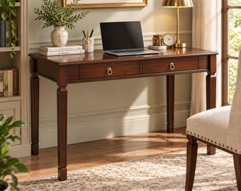 May include: A dark brown wooden writing desk featuring two drawers with gold ring pulls and fluted legs. The desk surface holds a silver laptop, a white ceramic vase with greenery, a marble pen holder, two stacked books, and a gold desk lamp.