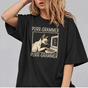 Può includere: Una t-shirt nera a maniche corte con la grafica di un gatto che digita su un computer. Il testo PURR-GRAMMER è scritto in beige chiaro sopra e sotto l'illustrazione. Il gatto è raffigurato in toni crema con accenti scuri.