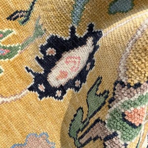 May include: A close-up view of a textured rug featuring a golden yellow background with intricate floral and botanical patterns in shades of navy blue, sage green, soft pink, and light blue. The woven pile shows a dense, knotted construction.