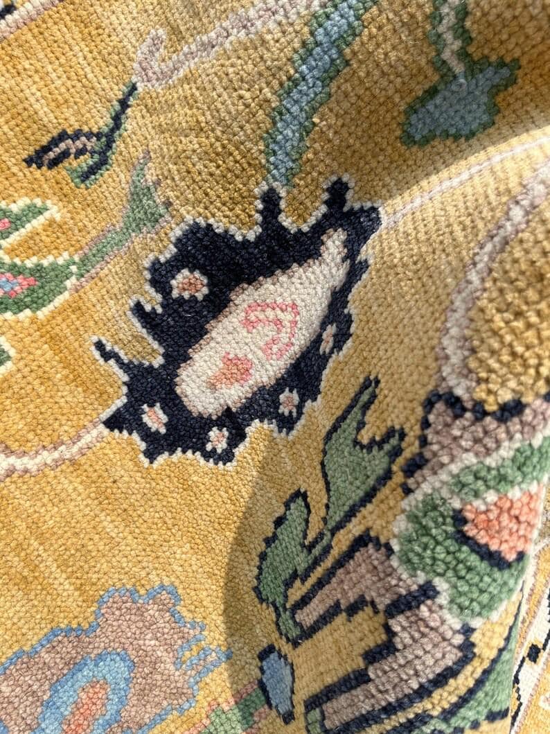 May include: A close-up view of a textured rug featuring a golden yellow background with intricate floral and botanical patterns in shades of navy blue, sage green, soft pink, and light blue. The woven pile shows a dense, knotted construction.