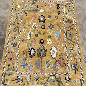 May include: A rectangular area rug featuring a mustard yellow background with a central field of stylized floral and botanical motifs in shades of blue, pink, green, white, and black. The rug has a decorative border and white fringe at both ends.