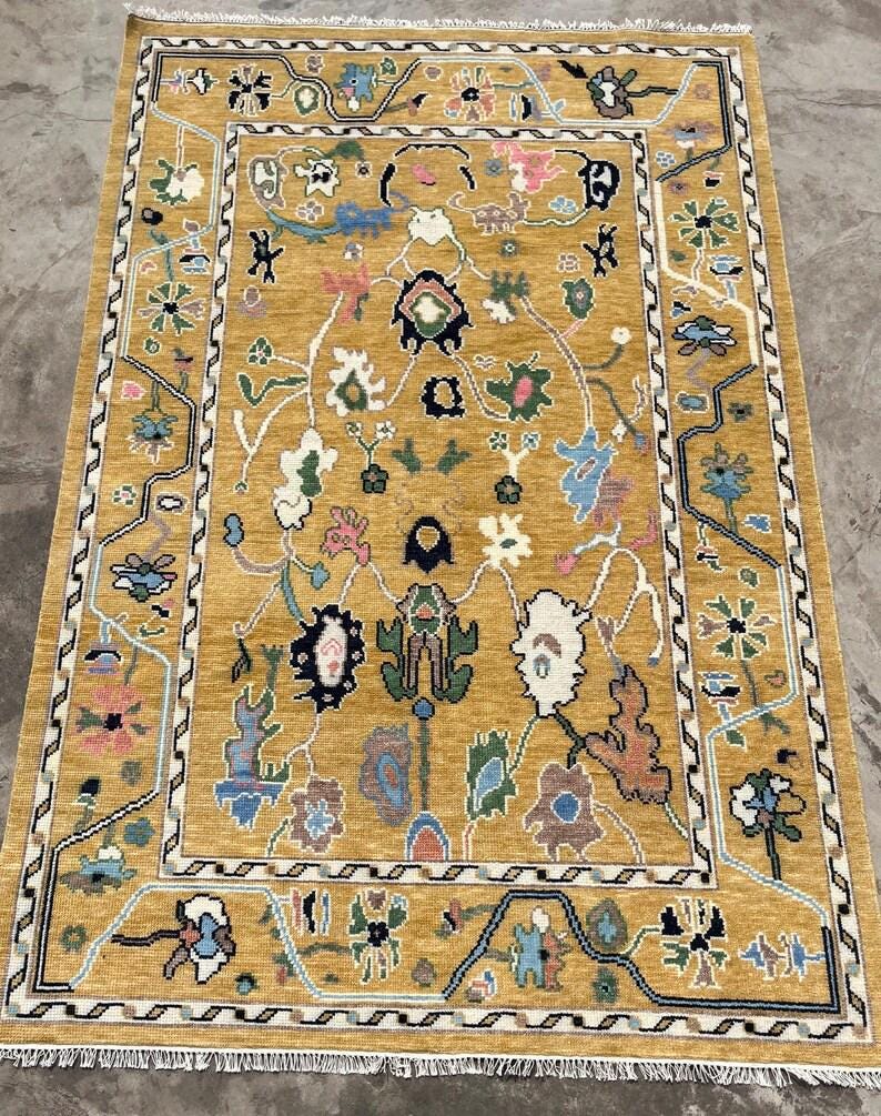 May include: A rectangular area rug featuring a mustard yellow background with a central field of stylized floral and botanical motifs in shades of blue, pink, green, white, and black. The rug has a decorative border and white fringe at both ends.