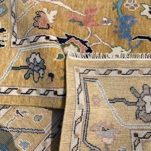 May include: A mustard yellow area rug featuring a detailed floral and geometric pattern in shades of blue, green, pink, and black. The rug has a visible woven texture, a decorative border, and a fringed edge, showing both the front and back sides.