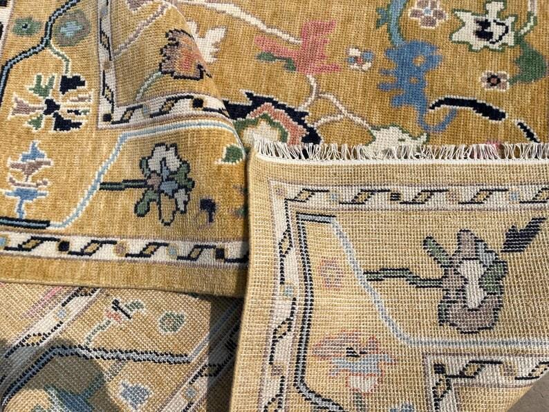 May include: A mustard yellow area rug featuring a detailed floral and geometric pattern in shades of blue, green, pink, and black. The rug has a visible woven texture, a decorative border, and a fringed edge, showing both the front and back sides.