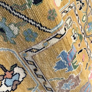 May include: A mustard yellow area rug featuring a dense, knotted pile texture. The surface displays a repeating geometric and floral pattern in shades of light blue, dark navy, white, and soft pink, organized within a structured border design.