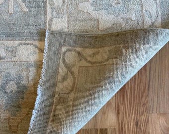 May include: A woven area rug featuring a light blue, cream, and beige floral pattern with a folded corner revealing the textured, light-colored underside. The rug displays a repeating geometric and botanical design across its surface.