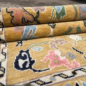May include: A mustard yellow area rug featuring a repeating floral and geometric pattern in shades of pink, light blue, dark green, and black. The rug is partially rolled, revealing a thick, textured pile and a detailed border with intricate motifs.