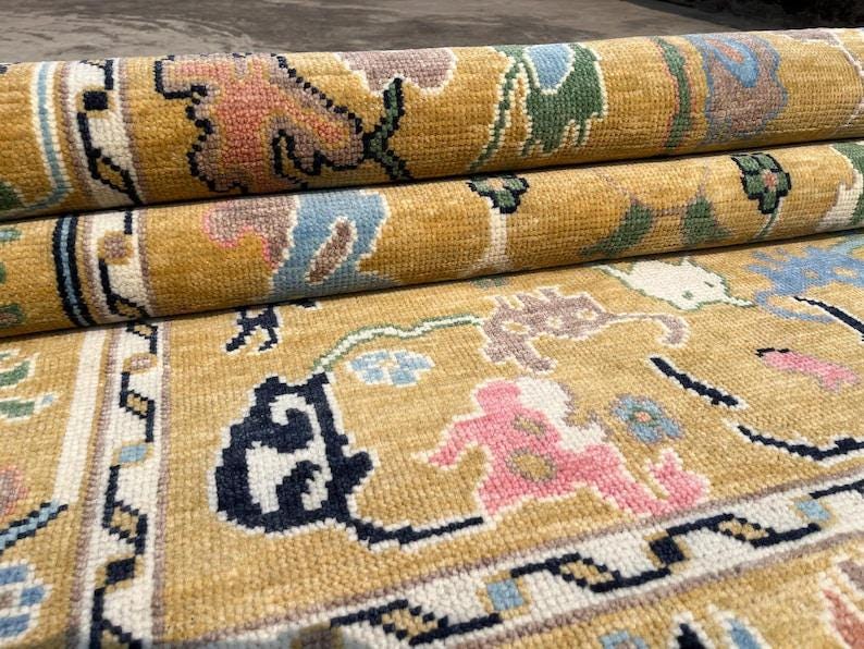 May include: A mustard yellow area rug featuring a repeating floral and geometric pattern in shades of pink, light blue, dark green, and black. The rug is partially rolled, revealing a thick, textured pile and a detailed border with intricate motifs.